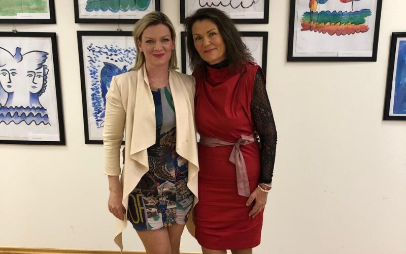 Barbi a Jitka z Ateliéru SZUŠ pózujú pred stenou s vystavenými farebnými výtvarnými dielami.